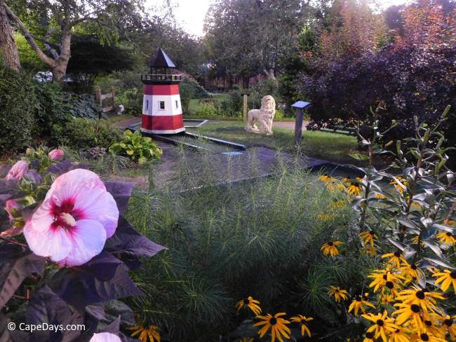 Mini golf course with blooming flowers, trees, a small lighthouse, and vintage stone lion statue.