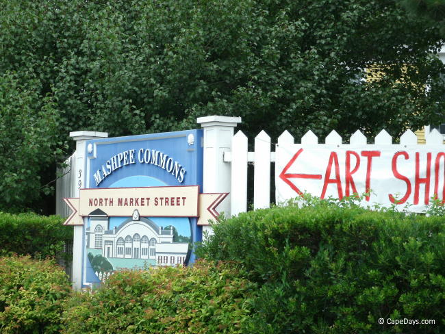 Green trees and shrubs surrounding a white picket fence with "Mashpee Commons" sign and banner advertising a weekend art show
