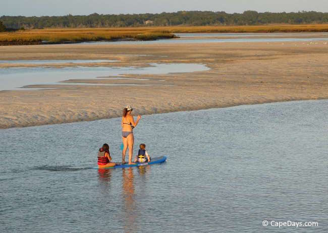 Woman paddleboarding with two young children on calm water in Yarmouth MA, with saltmarsh visible in the background.