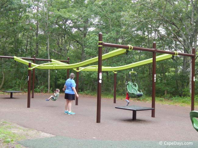 Woman watching her child ride the zip line at Bourne Inclusive Playground on Cape Cod, with trees in the background and another child pushing an empty zip line seat Woman watching her child ride the zip line at Bourne Inclusive Playground on Cape Cod, with trees in the background and another child pushing an empty zip line seat